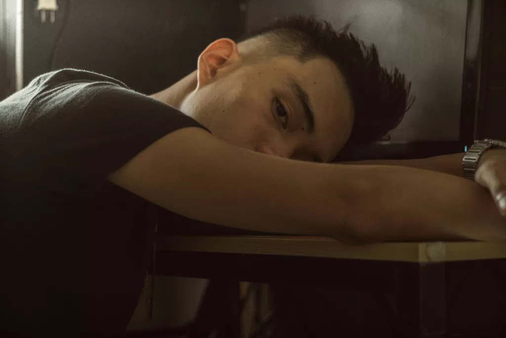 Serious male with modern haircut lying on table near monitor with head on arm and covering half face in room while looking at camera